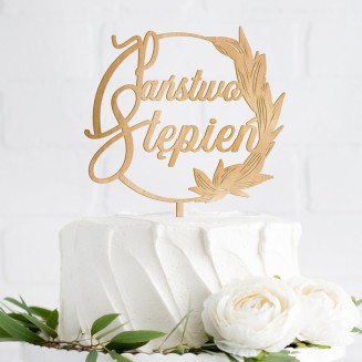 Topper na weselny tort. Wykonany z drewna. Ma kształt okręgu zdobionego z jednej strony listkami. W środku nazwisko nowożeńców.