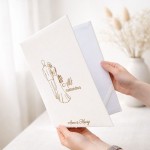 Personalizowana teczka na akt małżeństwa ma elegancką białą okładkę ze złotą ilustracją Pary Młodej i imionami