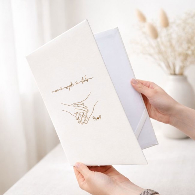 Personalizowana teczka na akt ślubu ma białą okładkę ze złotą grafiką splecionych dłoni oraz inicjałami Pary Młodej.
