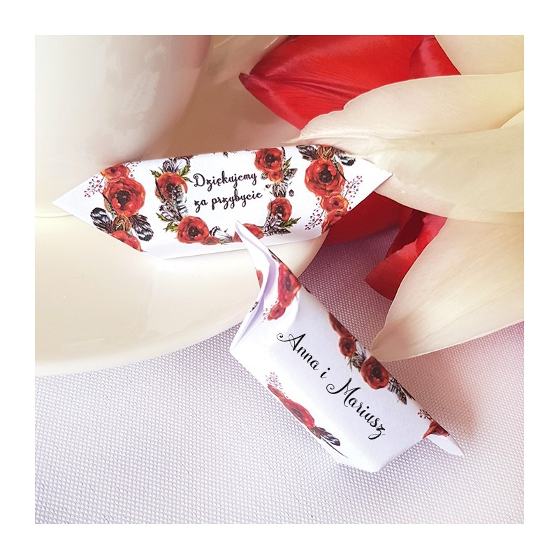 Dwie krówki w białych papierkach zdobionych nadrukiem czerwonych maków. Położone na beżowym talerzu. Z tyłu biało-kremowy kwiat.