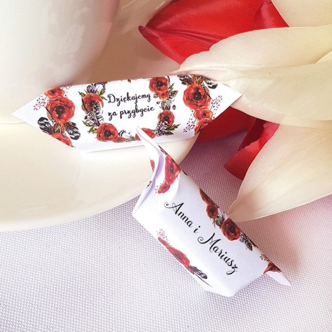 Dwie krówki w białych papierkach zdobionych nadrukiem czerwonych maków. Położone na beżowym talerzu. Z tyłu biało-kremowy kwiat.