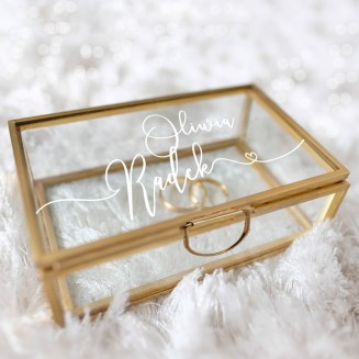 Szklana szkatułka na obrączki ze złotymi brzegami. Wieczko jest udekorowane białymi napisami.