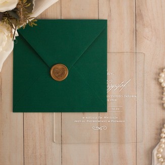 Akrylowe przezroczyste zaproszenie z tekstem zapisanym biała czcionką. Zielona koperta zamknięta złotym lakiem z motywem serca.